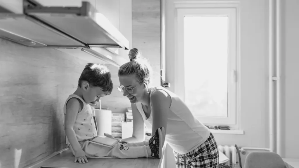 Parent smiling at a young child sitting on a kitchen counter, representing everyday learning, connection, and home education routines.