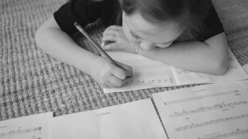 Child lying on the floor writing music notes in a workbook, representing creative learning in home education.