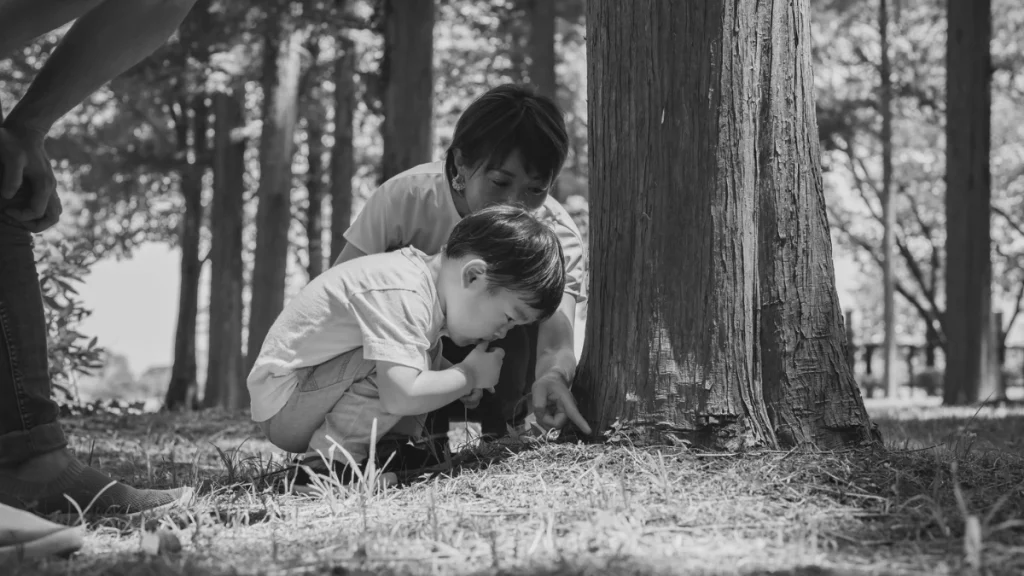 Parent supporting a young child as they explore the ground beside a tree, representing guided learning and curiosity in home education.