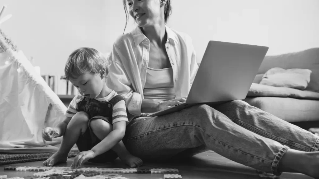 Parent using a laptop while a young child plays on the floor nearby, representing the challenge of balancing work, childcare, and home education.
