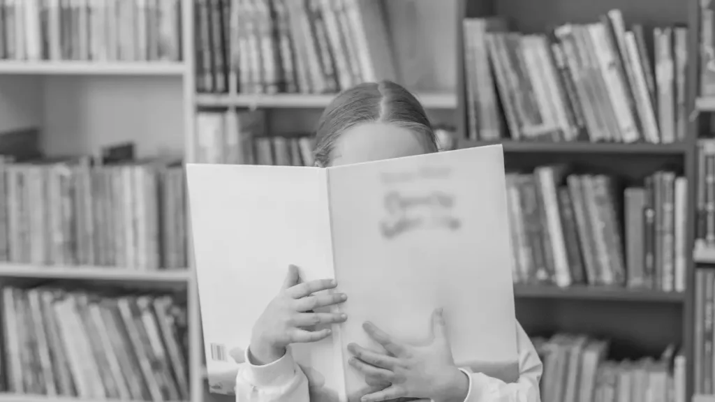 Child holding a book in front of a bookshelf, representing alternative learning routes, reading, and flexible education options.