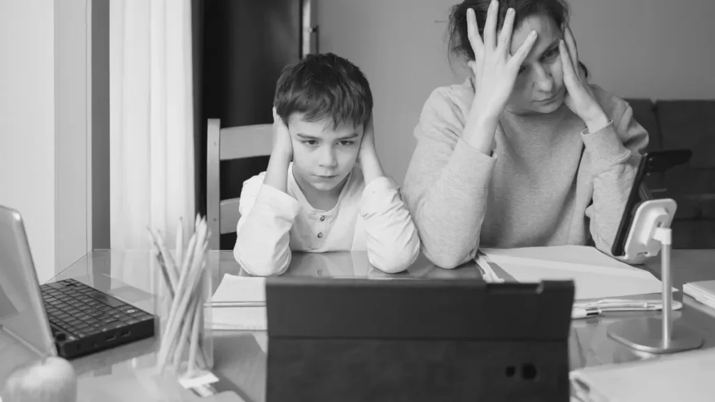 Parent and child looking stressed while working at a table with laptops and a phone, representing the misconception that home education is the same as lockdown learning.